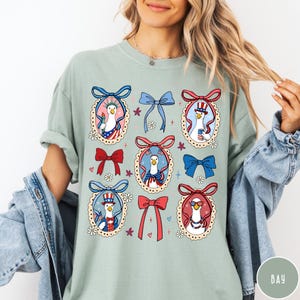 May include: Sage green t-shirt featuring a repeating pattern of patriotic geese in oval frames with bows. The geese are dressed in red, white, and blue, holding flags and wearing hats. Floral and heart accents complete the design.