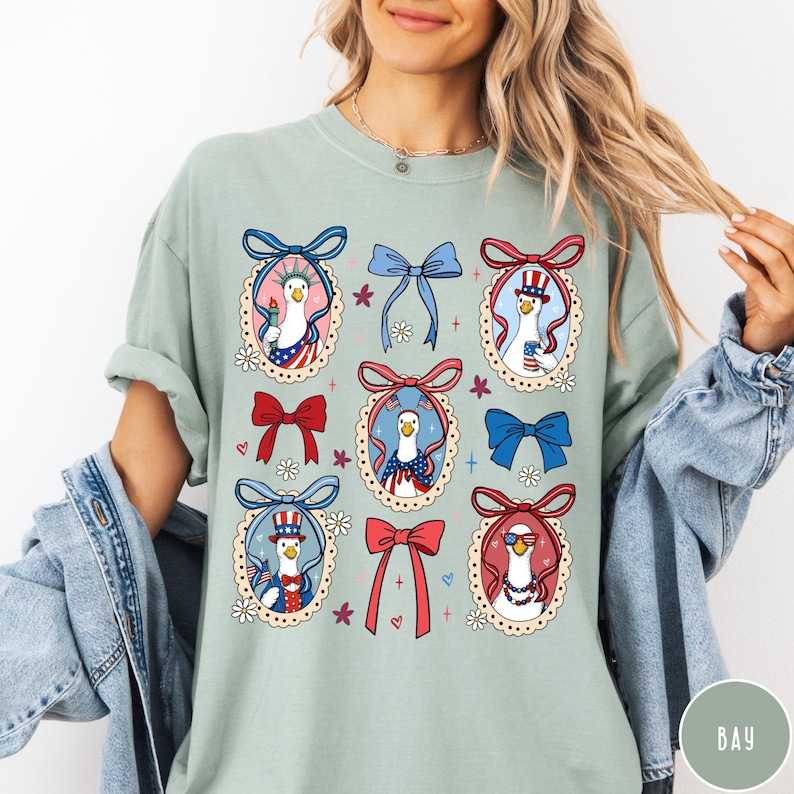 May include: Sage green t-shirt featuring a repeating pattern of patriotic geese in oval frames with bows. The geese are dressed in red, white, and blue, holding flags and wearing hats. Floral and heart accents complete the design.