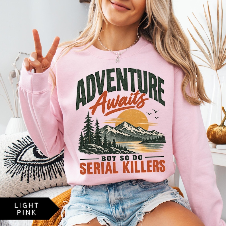 May include: A light pink crewneck sweatshirt featuring a graphic print with the text ADVENTURE Awaits BUT SO DO SERIAL KILLERS. The design includes a mountain landscape, a large sun, pine trees, and birds in dark green, orange, and black ink.