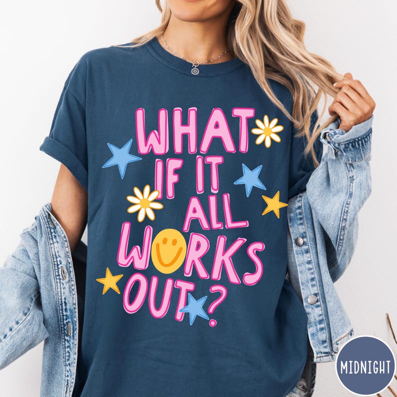 May include: A dark blue short-sleeve t-shirt featuring the pink text "WHAT IF IT ALL WORKS OUT?" with a yellow smiley face replacing the letter "O". The design includes scattered white daisies, yellow stars, and light blue stars across the front.