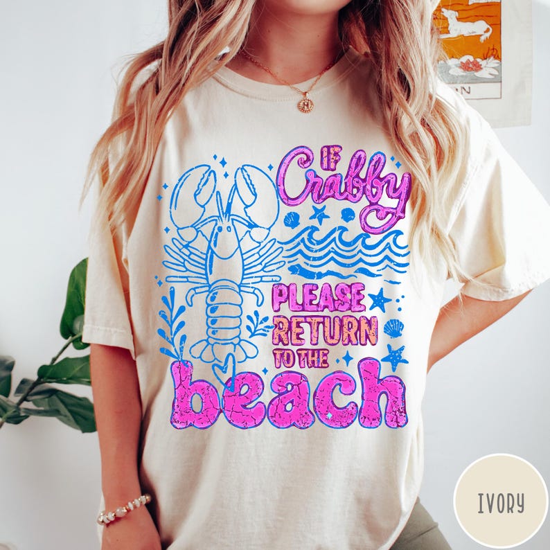 Puede incluir: Camiseta color marfil con un dise&ntilde;o gr&aacute;fico desgastado. El dise&ntilde;o presenta una langosta azul, olas y el texto "If Crabby Please Return to the Beach" en rosa y azul. La camiseta es de corte holgado.