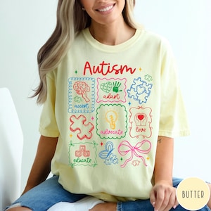 May include: Butter-colored t-shirt with the word "Autism" in red script. The shirt features colorful illustrations and words like "accept", "adapt", "advocate", "love", and "educate". The design includes puzzle pieces, hearts, and a brain.