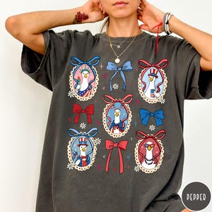 May include: Dark gray t-shirt featuring a repeating pattern of cartoon geese in patriotic outfits, framed by decorative bows and floral accents. The geese are depicted with American flag motifs and accessories, creating a festive design.