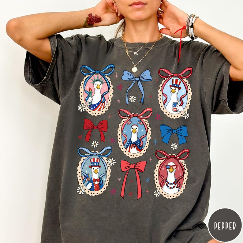 May include: Dark gray t-shirt featuring a repeating pattern of cartoon geese in patriotic outfits, framed by decorative bows and floral accents. The geese are depicted with American flag motifs and accessories, creating a festive design.