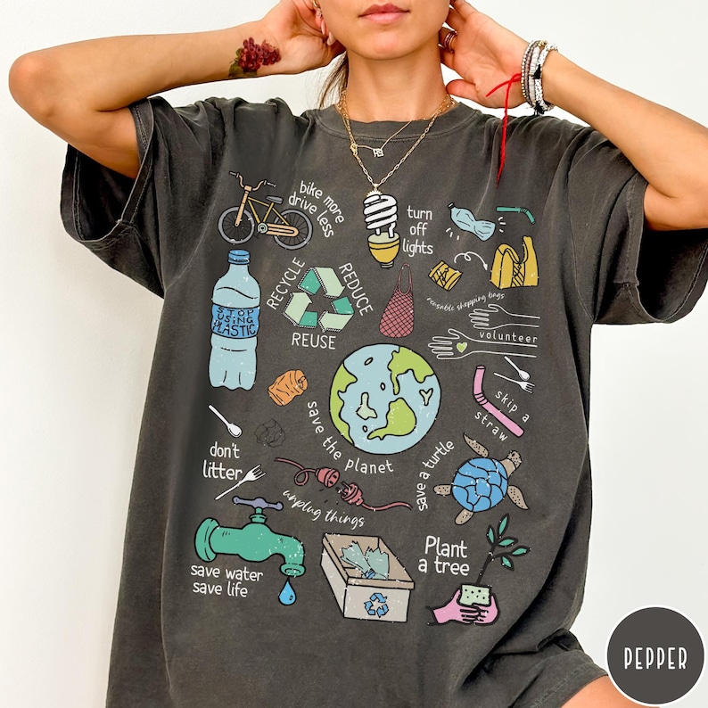 May include: A dark grey t-shirt featuring a graphic print of various illustrations related to environmentalism. The illustrations include a bicycle, a lightbulb, a turtle, a plant, a recycling symbol, and the text "bike more drive less", "turn off lights", "reduce", "recycle", "reuse", "volunteer", "skip a straw", "don't litter", "save the planet", "save a turtle", "unplug things", "save water save life", and "plant a tree".
