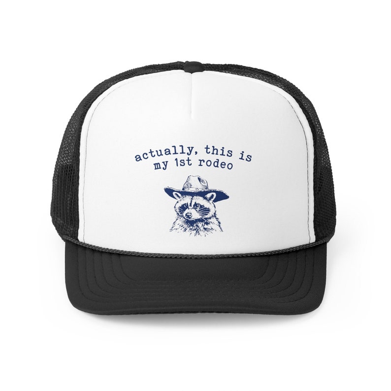 May include: A black and white trucker hat with a white front panel. The front panel features a raccoon wearing a cowboy hat and the text "actually, this is my 1st rodeo".