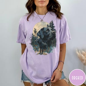 May include: A light purple t-shirt featuring a graphic of a black Labrador Retriever with a full moon and forest scene. The word "ORCHID" is printed on a pink circle in the lower right corner. The shirt has short sleeves.