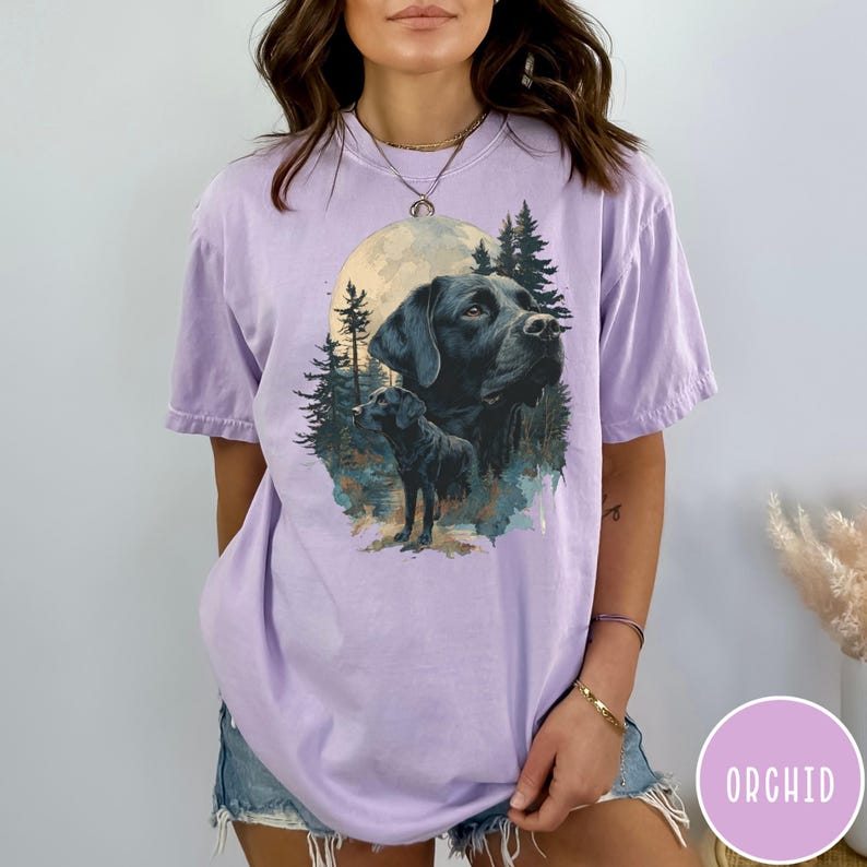May include: A light purple t-shirt featuring a graphic of a black Labrador Retriever with a full moon and forest scene. The word "ORCHID" is printed on a pink circle in the lower right corner. The shirt has short sleeves.