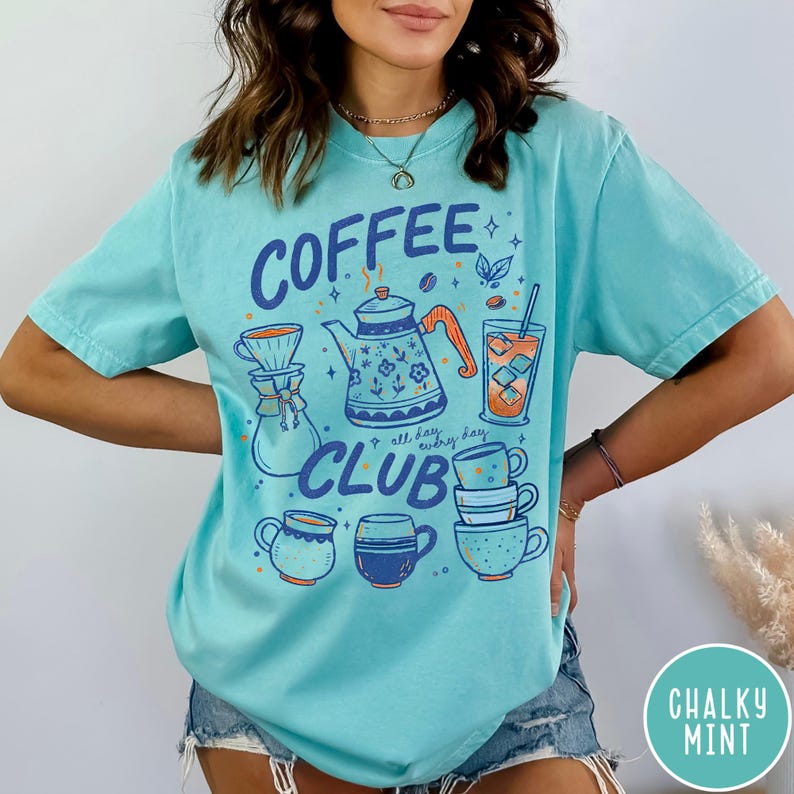 May include: A light turquoise t-shirt with a "Coffee Club" graphic. The design includes a coffee pot, cups, and iced coffee. The text "all day, everyday" is also included in the design. The shirt is a comfortable, casual style.