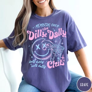 May include: A purple t-shirt with a graphic design featuring the text "MEMBERS ONLY the Dilly Dally Club." The design includes a smiley face, a drink, and the words "half dilly, half dally."