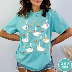 K&ouml;nnte beinhalten: Ein t&uuml;rkisfarbenes T-Shirt mit einem Motiv von wei&szlig;en G&auml;nsen, die bunte Schleifen tragen. Die G&auml;nse sind von kleinen Blumenmustern umgeben.