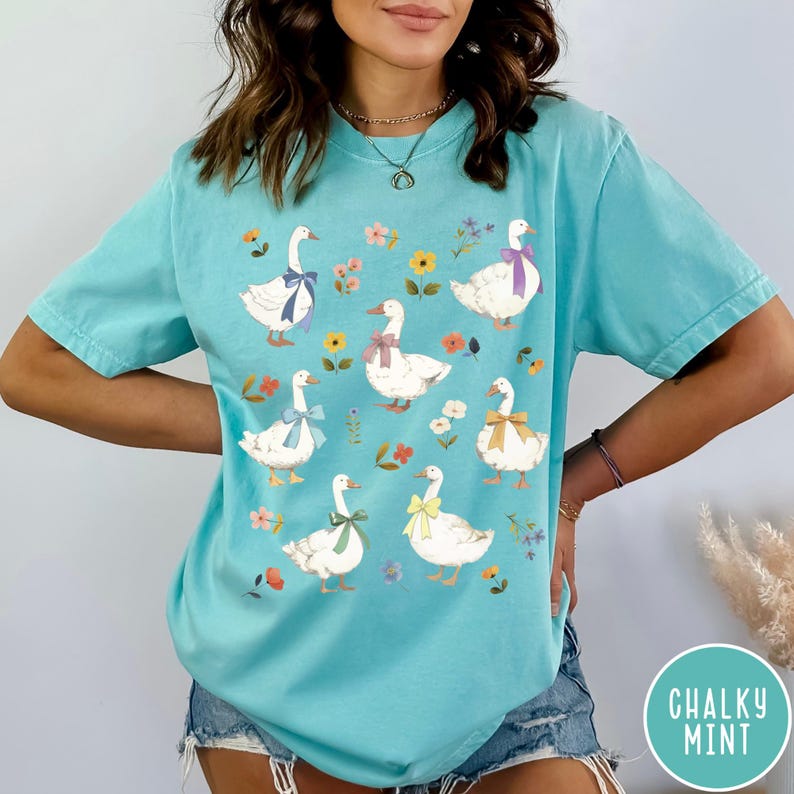 K&ouml;nnte beinhalten: Ein t&uuml;rkisfarbenes T-Shirt mit einem Motiv von wei&szlig;en G&auml;nsen, die bunte Schleifen tragen. Die G&auml;nse sind von kleinen Blumenmustern umgeben.