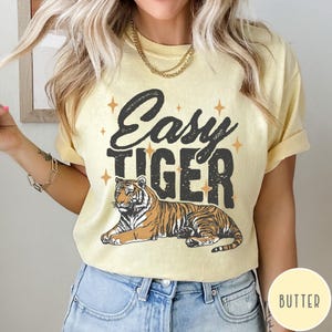 May include: A yellow t-shirt with the text "Easy Tiger" in black lettering and a stylised tiger illustration. The shirt has a distressed look and features small gold stars.