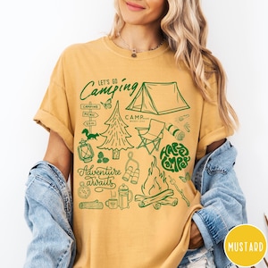 Może przedstawiać: Musztardowa koszulka z zieloną grafiką o tematyce kempingowej. Projekt zawiera namiot, ognisko, drzewa oraz napisy "Let's Go Camping", "Adventure Awaits" i "Happy Campers".