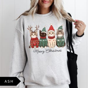 May include: Light gray sweatshirt featuring a graphic of four cats dressed in Christmas sweaters and hats. The cats are wearing red, green, and white outfits. The text "Meowy Christmas" is written below the cats.