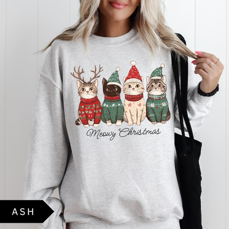 May include: Light gray sweatshirt featuring a graphic of four cats dressed in Christmas sweaters and hats. The cats are wearing red, green, and white outfits. The text "Meowy Christmas" is written below the cats.