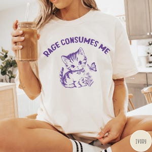 Puede incluir: Camiseta blanca con un gr&aacute;fico morado de un gato de dibujos animados con un lazo y el texto "Rage Consumes Me".