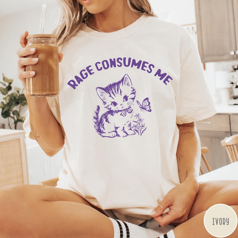 Puede incluir: Camiseta blanca con un gr&aacute;fico morado de un gato de dibujos animados con un lazo y el texto "Rage Consumes Me".