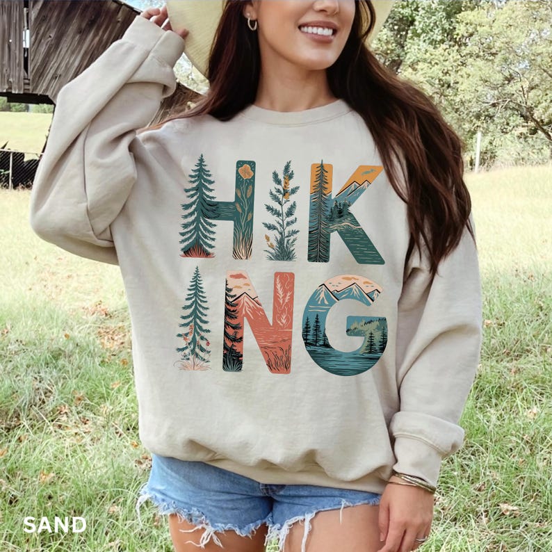 Puede incluir: Sudadera de color arena con la palabra "HIKING" en letras coloridas con tem&aacute;tica de la naturaleza. Las letras est&aacute;n dise&ntilde;adas con im&aacute;genes de &aacute;rboles, monta&ntilde;as y paisajes. La sudadera es de color neutro y tiene mangas largas.