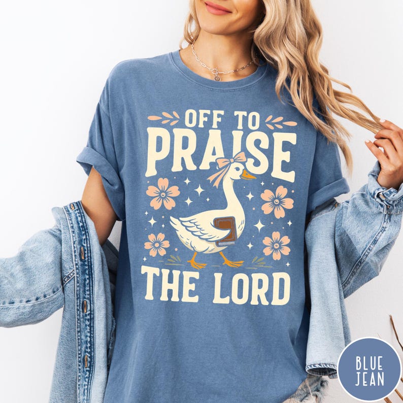 Puede incluir: Camiseta azul con el texto "OFF TO PRAISE THE LORD" y un dibujo de un ganso con lazo y libro. Flores y estrellas color melocot&oacute;n rodean al ganso. La camiseta es de color azul liso.