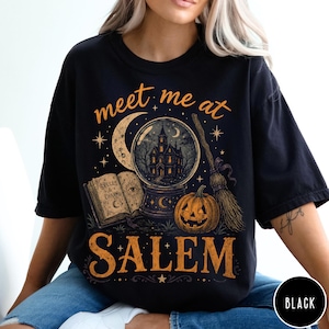 May include: A black t-shirt featuring a graphic print with the text "meet me at SALEM" in orange. The design includes a crystal ball containing a haunted house, an open spellbook, a crescent moon, stars, a jack-o'-lantern, and a broomstick.