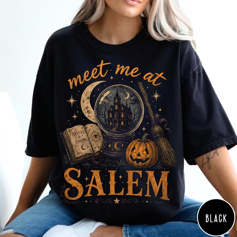 May include: A black t-shirt featuring a graphic print with the text "meet me at SALEM" in orange. The design includes a crystal ball containing a haunted house, an open spellbook, a crescent moon, stars, a jack-o'-lantern, and a broomstick.
