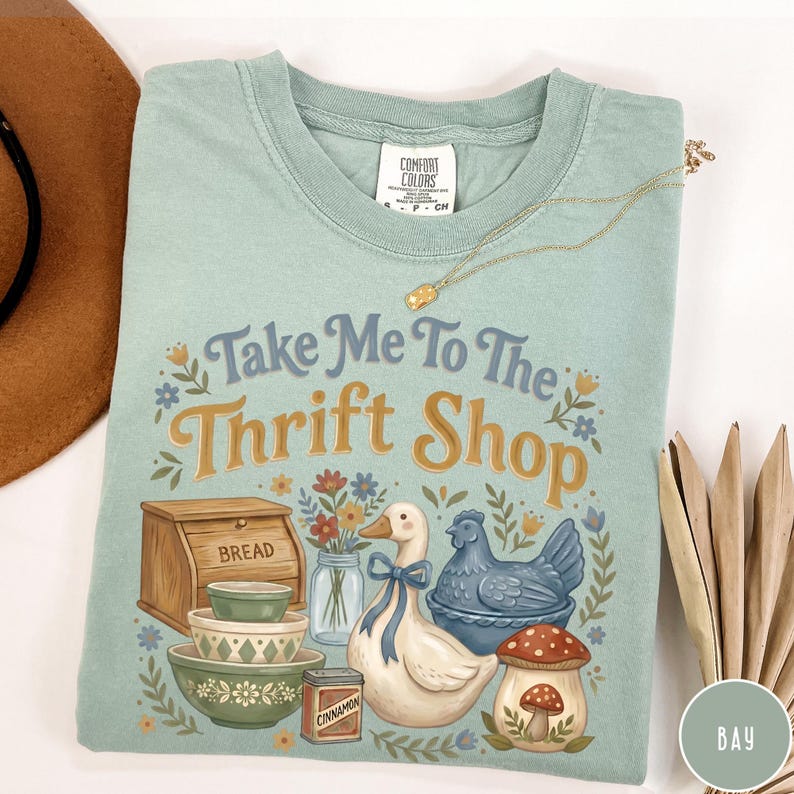 Op de afbeelding: Een lichtgroen Comfort Colors T-shirt met de tekst "Take Me To The Thrift Shop" en illustraties van een brooddoos, kommen, een eend, een kip en een paddenstoel. Een gouden ketting ligt over het shirt.