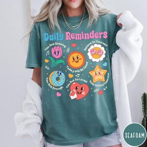 May include: Teal t-shirt with the words "Daily Reminders" in pink and blue. The shirt features cartoon graphics of a flower, sun, donut, star, and heart, each with positive affirmations. The shirt is a comfortable, casual style.