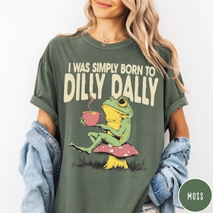 May include: Moss green t-shirt with the text "I WAS SIMPLY BORN TO DILLY DALLY". The graphic features a cartoon frog holding a pink teacup while sitting on a mushroom. The shirt is a relaxed fit.