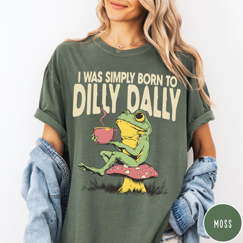 May include: Moss green t-shirt with the text "I WAS SIMPLY BORN TO DILLY DALLY". The graphic features a cartoon frog holding a pink teacup while sitting on a mushroom. The shirt is a relaxed fit.