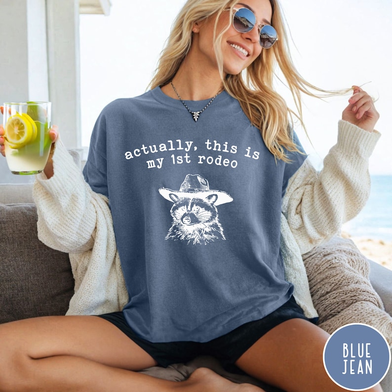 May include: A blue jean t-shirt with a white graphic of a raccoon wearing a cowboy hat. The text "actually, this is my 1st rodeo" is printed on the shirt.