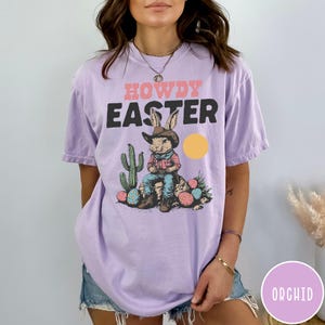 Puede incluir: Una camiseta de color lila con las palabras "HOWDY EASTER" en rosa y negro. El gr&aacute;fico presenta un conejo vaquero con un cactus, huevos de Pascua y un sol amarillo. La camiseta es de estilo casual y unisex.