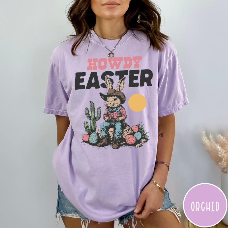 Puede incluir: Una camiseta de color lila con las palabras "HOWDY EASTER" en rosa y negro. El gr&aacute;fico presenta un conejo vaquero con un cactus, huevos de Pascua y un sol amarillo. La camiseta es de estilo casual y unisex.
