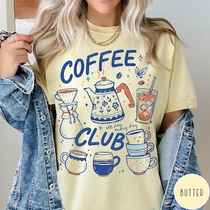 May include: A light yellow t-shirt with a blue and orange graphic design featuring coffee-related illustrations and the words "COFFEE CLUB" and "all day, every day". The design includes a teapot, coffee cups, and a glass of iced coffee.