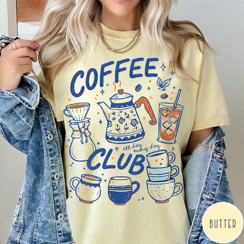 May include: A light yellow t-shirt with a blue and orange graphic design featuring coffee-related illustrations and the words "COFFEE CLUB" and "all day, every day". The design includes a teapot, coffee cups, and a glass of iced coffee.