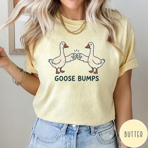 Puede incluir: Una camiseta de manga corta de color amarillo p&aacute;lido con un gr&aacute;fico de dos gansos blancos d&aacute;ndose un choque de pu&ntilde;os. Debajo de los gansos, el texto GOOSE BUMPS est&aacute; impreso en letras may&uacute;sculas de color azul oscuro. Tiene cuello redondo.