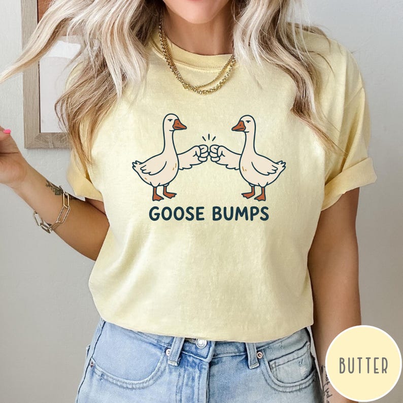 Puede incluir: Una camiseta de manga corta de color amarillo p&aacute;lido con un gr&aacute;fico de dos gansos blancos d&aacute;ndose un choque de pu&ntilde;os. Debajo de los gansos, el texto GOOSE BUMPS est&aacute; impreso en letras may&uacute;sculas de color azul oscuro. Tiene cuello redondo.