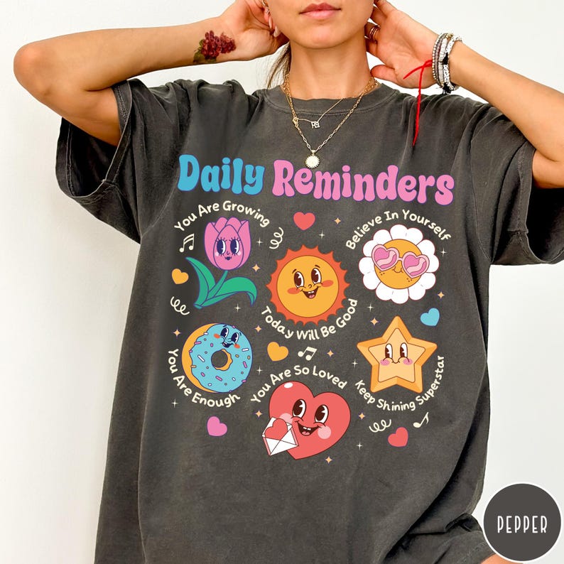 May include: Dark gray t-shirt with the text "Daily Reminders" in pink and blue. The shirt features colorful cartoon graphics of a flower, sun, donut, star, and heart with positive affirmations. The shirt is a casual, comfortable style.