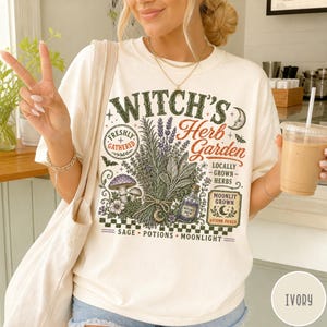 May include: An ivory t-shirt featuring a graphic print with the text "Witch's Herb Garden". The design includes illustrations of a bundle of herbs, mushrooms, a potion bottle, a crescent moon, and the words "Freshly Gathered", "Locally Grown Herbs", "Moonlit Grown", "Autumn Picked", "Sage", "Potions", and "Moonlight".