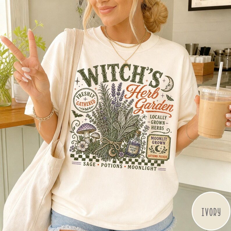May include: An ivory t-shirt featuring a graphic print with the text "Witch's Herb Garden". The design includes illustrations of a bundle of herbs, mushrooms, a potion bottle, a crescent moon, and the words "Freshly Gathered", "Locally Grown Herbs", "Moonlit Grown", "Autumn Picked", "Sage", "Potions", and "Moonlight".