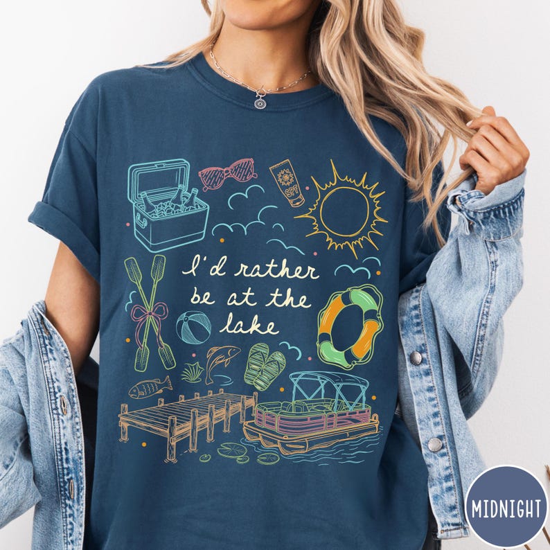 Puede incluir: Camiseta azul marino con un gr&aacute;fico caprichoso con tem&aacute;tica de lago. El dise&ntilde;o incluye una nevera, gafas de sol, protector solar, un sol, nubes, remos, un bal&oacute;n de playa, un pez, sandalias, un muelle, un salvavidas y un barco. El texto dice "I'd rather be at the lake."