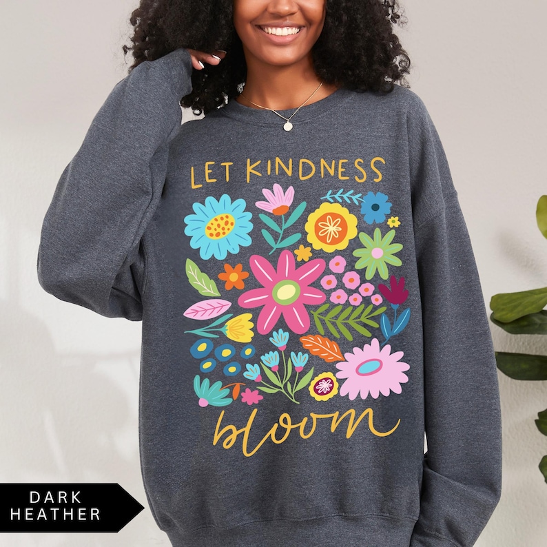 May include: A dark heather grey sweatshirt with a colorful floral design and the text "Let Kindness Bloom" in yellow.