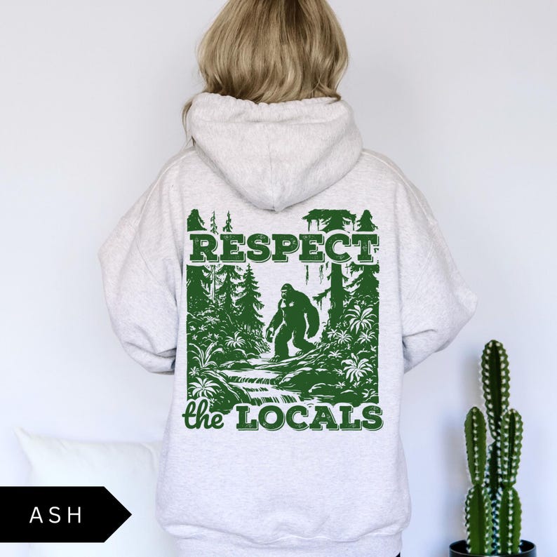 May include: A gray hooded sweatshirt with a green graphic print of a Bigfoot walking through a forest. The graphic says "Respect the Locals".