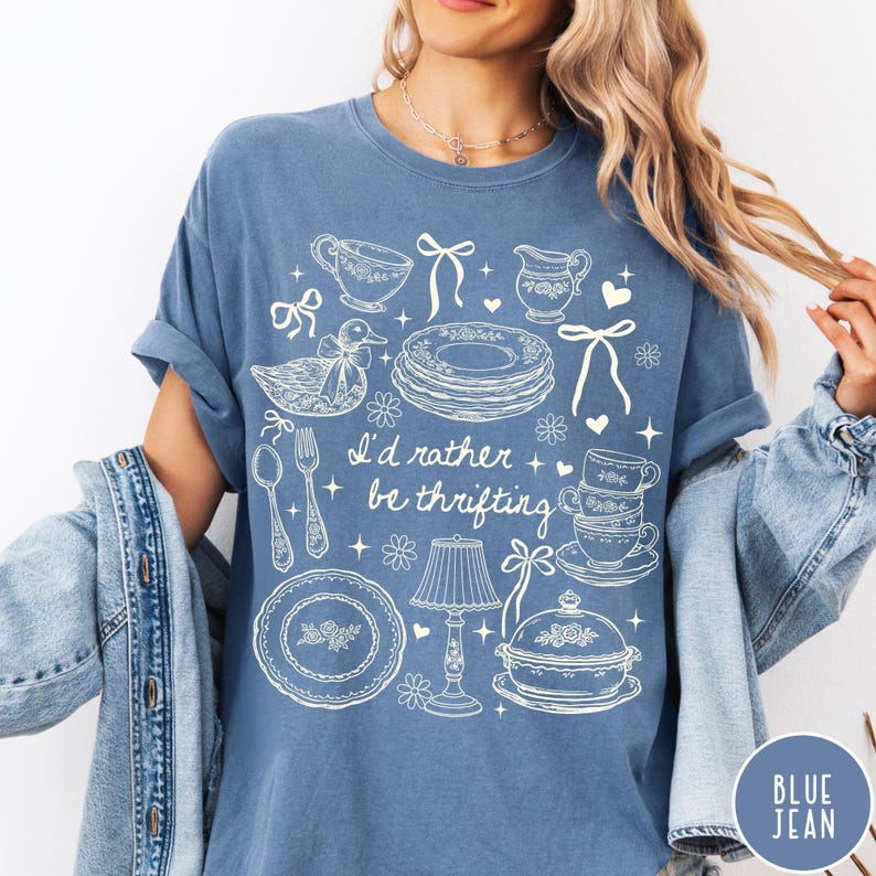 May include: Blue jean-coloured t-shirt with a white graphic design featuring teacups, plates, and the text "I'd rather be thrifting." The design includes a duck, bows, and a lamp. The shirt has short sleeves and a relaxed fit.