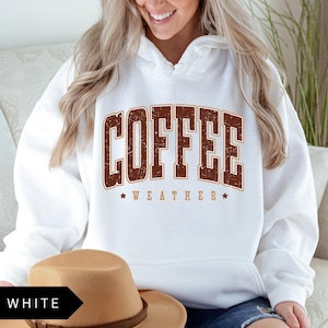 Puede incluir: Sudadera blanca con un gr&aacute;fico marr&oacute;n de estilo vintage que dice "Coffee Weather" con una estrella a cada lado del texto.