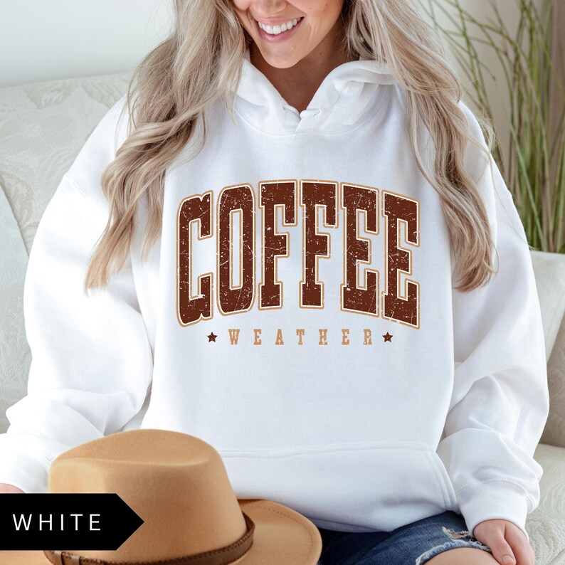 Puede incluir: Sudadera blanca con un gr&aacute;fico marr&oacute;n de estilo vintage que dice "Coffee Weather" con una estrella a cada lado del texto.