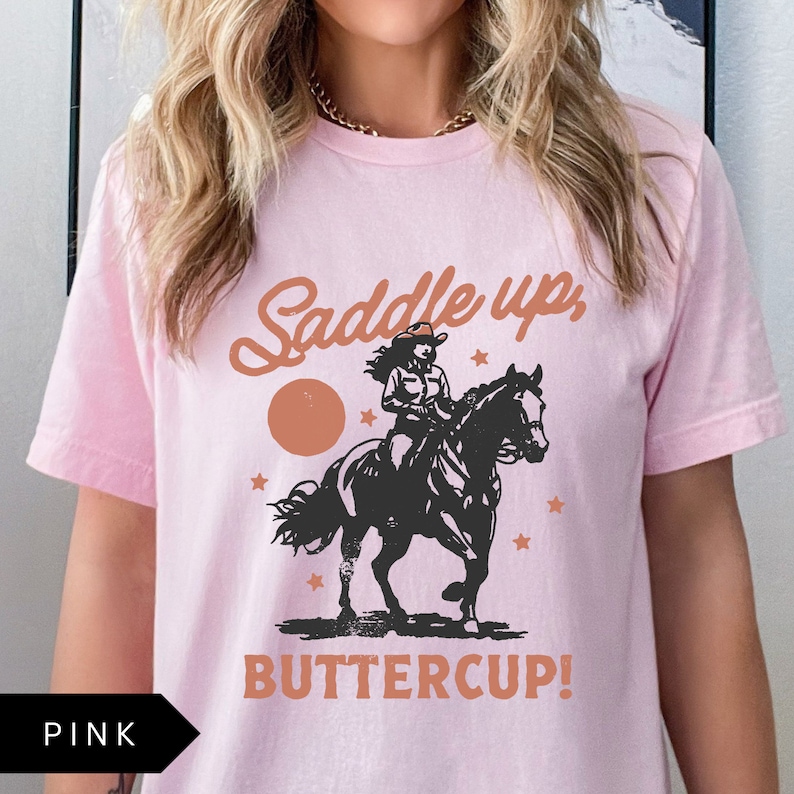 May include: Pink t-shirt with a graphic of a woman riding a horse with the text "Saddle up, Buttercup!"