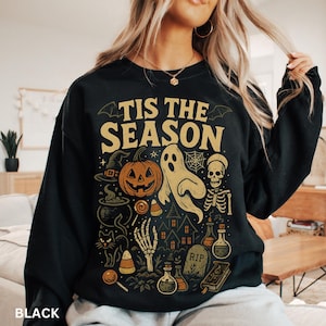 Puede incluir: Sudadera negra con un dise&ntilde;o de Halloween. El dise&ntilde;o incluye una calabaza, un fantasma, un esqueleto y el texto "TIS THE SEASON" en letras doradas. Otros elementos incluyen un sombrero de bruja, un caldero y caramelos.