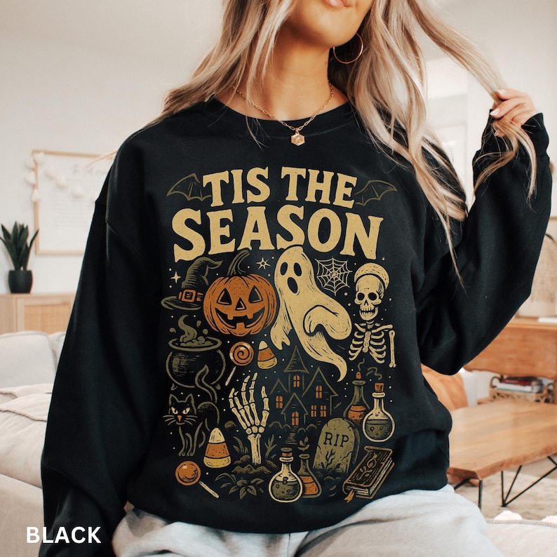 Puede incluir: Sudadera negra con un dise&ntilde;o de Halloween. El dise&ntilde;o incluye una calabaza, un fantasma, un esqueleto y el texto "TIS THE SEASON" en letras doradas. Otros elementos incluyen un sombrero de bruja, un caldero y caramelos.
