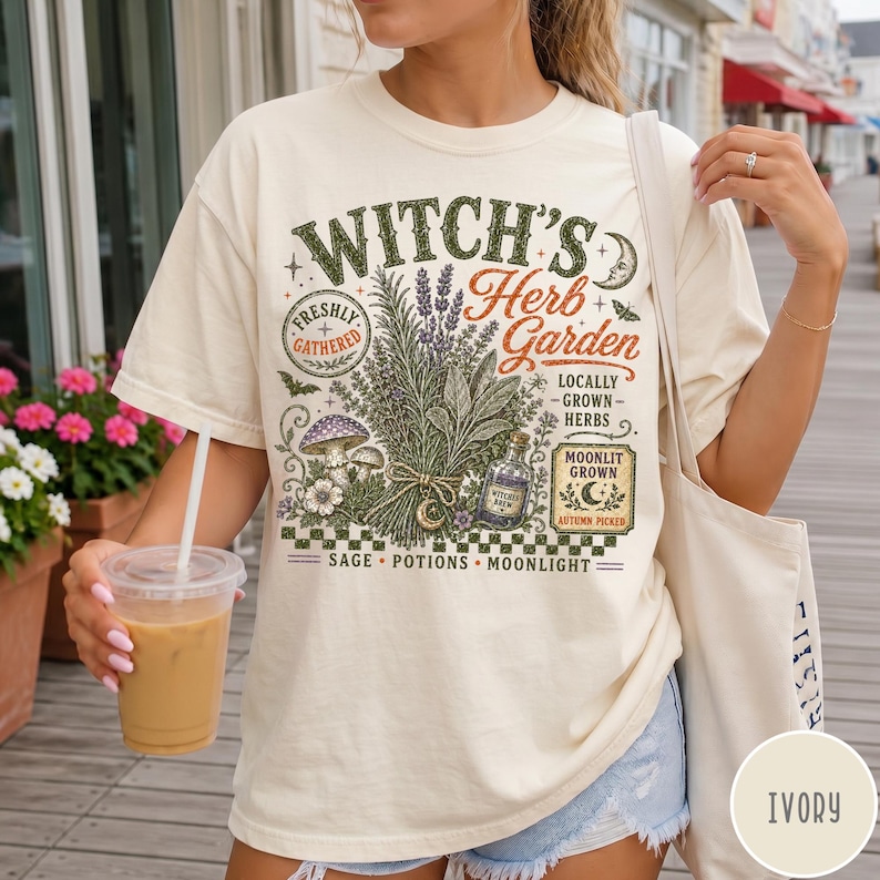May include: An ivory t-shirt featuring a graphic print with the text "Witch's Herb Garden," "Freshly Gathered," "Locally Grown Herbs," "Moonlit Grown," "Autumn Picked," and "Sage, Potions, Moonlight." The design includes illustrations of herbs, mushrooms, and a bottle.